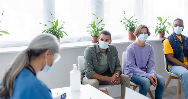 Auf dem Foto ist links eine Krankenpflegerin in Arbeitskleidung zu sehen, die an einem Tisch sitzt und etwas schreibt. Rechts im Bild sitzen zwei Männer und eine Frau auf Stühlen. Sie schauen zur Pflegerin, ihre Blicke und ihre Körperhaltungen wirken nervös.