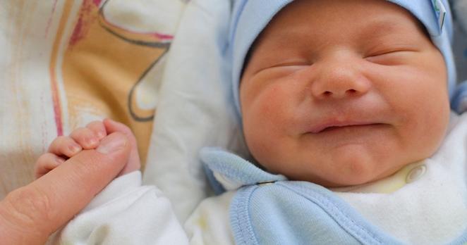 Ein neugeborenes Baby mit geschlossenen Augen und einer blauen Mütze umfasst den Zeigefinger eines Erwachsenen.