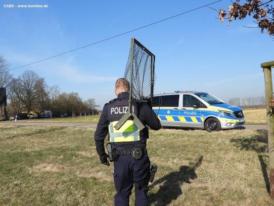 Auf dem Bild ist ein Polizist zu sehen, der eine große Vogelfallen auf den Schultern zu einem Streifenwagen trägt 