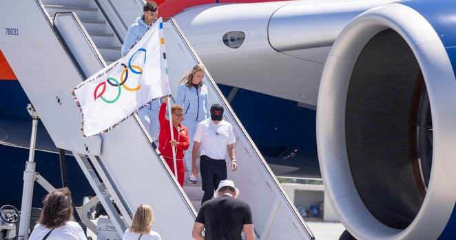 Bürgrmeisterin Bass in rotem Frottee-Trainingsanzug geht die Treppe vom Flugzeug aufs Rollfeld hinunter. Sie hält die Flagge. Auf dem Rollfeld wartet ein kleines Empfangskomittee.