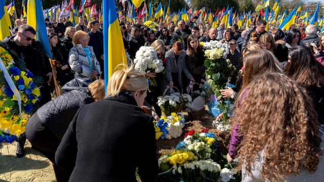 Auf einem Friedhof legen Menschen Blumen auf einem Grab nieder. Viele Trauergäste haben ukrainische Flaggen in der Hand.