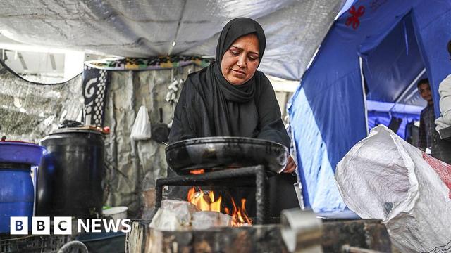 A woman wearing a head covering cooks on a makeshift stove in a tent in Gaza City