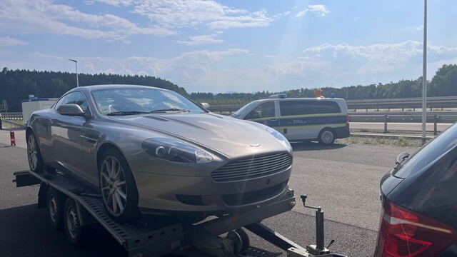 Ein silberner Aston Martin ist auf einem Fahrzeuganhänger verladen. Im Hintergrund steht ein Einsatzfahrzeug des Zolls.