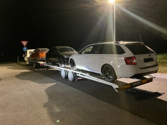 Ein weißer Transporter mit Fahrzeuganhänger hat ein orangenes, ein schwarzes und ein weißes Auto geladen. Es ist Nacht.