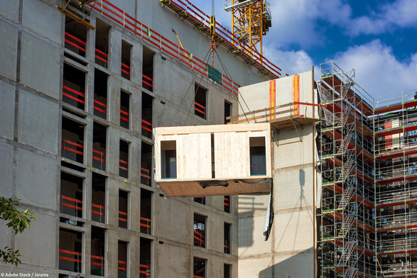 Ein Holzmodul an einem Kranseil vor einem im Bau befindlichen Gebäude.