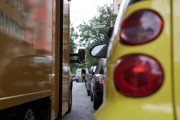 Ein Foto, das eine Straßensituation zeigt: Zu sehen ist rechts im Bild ein parkendes Auto, das offenbar auf einer Straße gemeinsam mit vielen anderen Autos in Längs-Parkrichtung parkt. Links im Bild steht auf der Fahrbahn, neben den parkenden Autos in zweiter Reihe, ein Lieferfahrzeug in gelber Farbe. Ein paar Meter vor diesem (Post-)Lieferfahrzeug steht ein weiteres Lieferauto in zweiter Reihe, bei welchem gerade etwas ausgeladen wird.