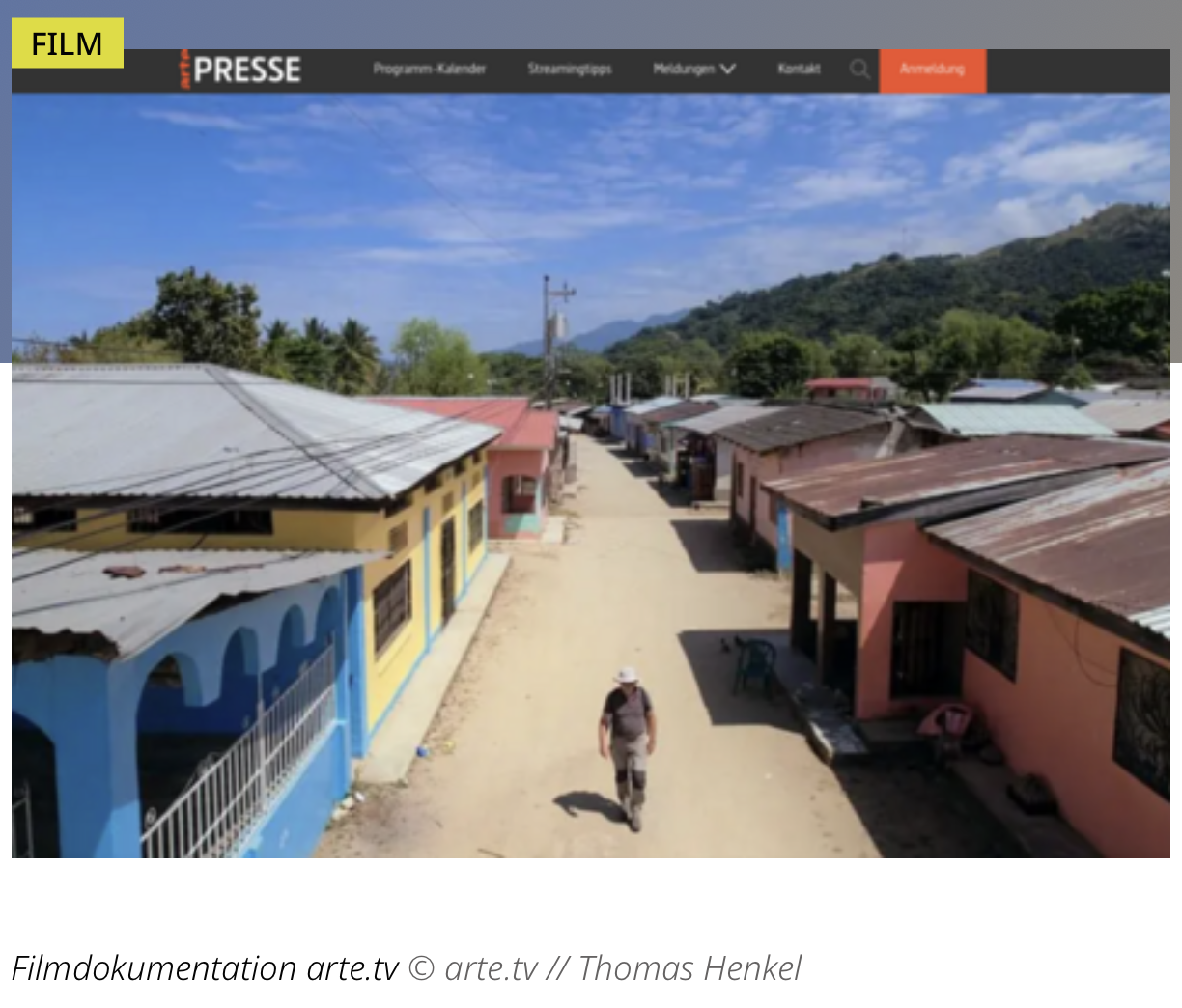 Screenshot aus dem "Honduras"-Dokumentarfilm, der eine unbefestigte Straße gesäumt von flachen, in verschiedenen Farben gestrichenen Häusern zeigt. Ein Mann läuft die Straße entlang, im Hintergrund sind dicht bewaldete Berge zu sehen.