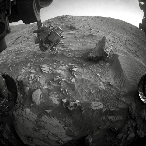 Die selbe Szene aus Sicht von einer der Bordkameras. Links ragt der mechanische Arm des Rovers ins Bild. An seinem Kopf befinden sich mehrere Instrumente und eben auch die Kamera, die das vorher gezeigte Foto aufnahm. Sie befindet sich hier nur knapp über dem sandigen und von Gestein übersäten Boden.
