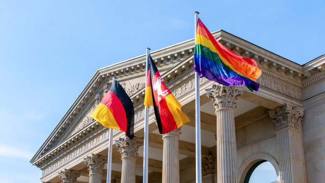 Foto des oberen Teil des Portikus, davor wehen von links nach rechts die Deutschlandflagge, die Niedersachsenflagge die Regenbogenflagge.