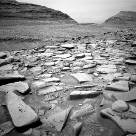Vorne eine flache Oberfläche voller Steinplatten, die dort lose herumliegen. Im Hintergrund eine offenbar eher sandige Landschaft und zwei runde Hügel.