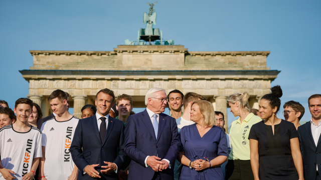 Bundesinnen- und Sportministerin Nancy Faeser, Bundespräsident Frank-Walter Steinmeier und der französische Präsident Emmanuel Macron stehen am Brandenburger Tor in Berlin.
Sie sind umringt um weiteren Personen.