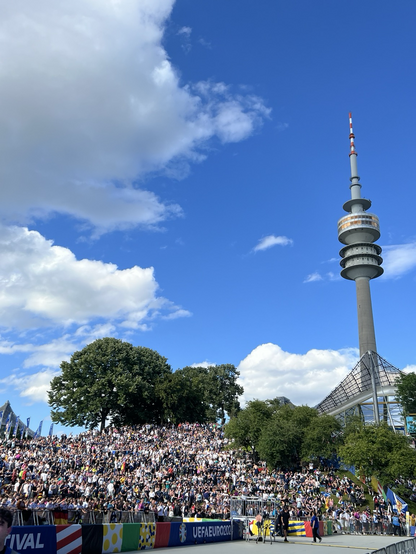 Blick in den Olympiapark München mit Olympiaturm. Viele Fußballfans sitzen und stehen auf den Hügeln vor der Bühne.
