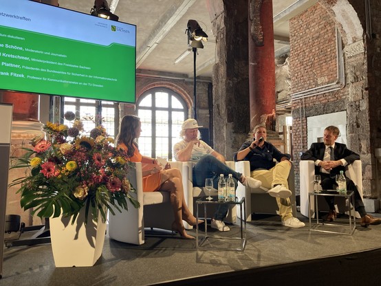 BSI-Präsidentin Claudia Plattner sitzt beim Frauennetzwerktreffen in Dresden auf einem Panel u.a. neben dem sächsischen Ministerpräsidenten Michael Kretschmer und hält ein Mikrofon in der Hand, in das sie spricht.