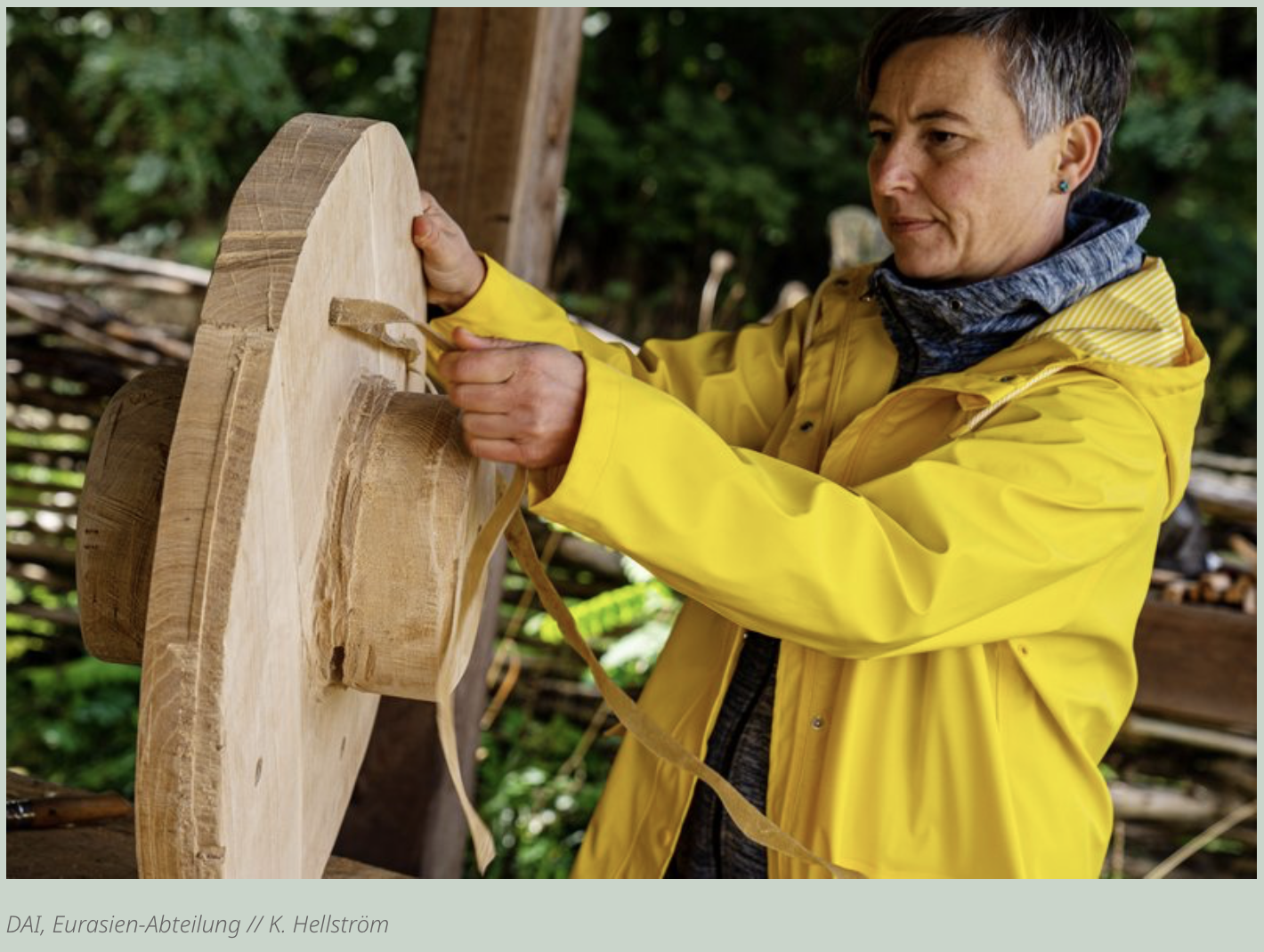 Foto einer Frau mit kurzen dunklen Haaren in gelber Regenjacke, die mit einem Riemen an einem Holzrad hantiert.