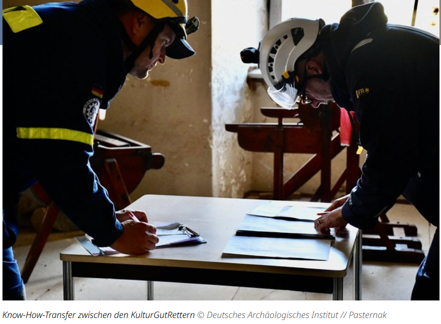 Foto, das zwei über einen in offenabr provisiorisch eingerichteten Raum stehenden Tisch gebeute Personen zeigt. Beide tragen Schutzhelme und THW-Einsatz- bzw. Outdoorkleidung. Auf dem Tisch liegen Unterlagen, mit denen sich beide Personen befassen.