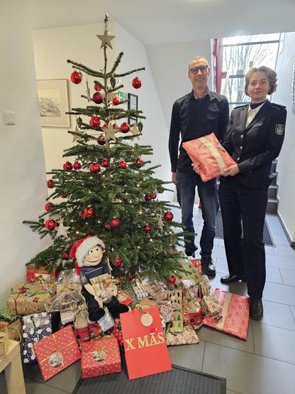 Zwei Beschäftigte des Zolls stehen neben einem geschmückten Weihnachtsbaum, unter dem zahlreiche Geschenke für benachteiligte Kinder liegen. 