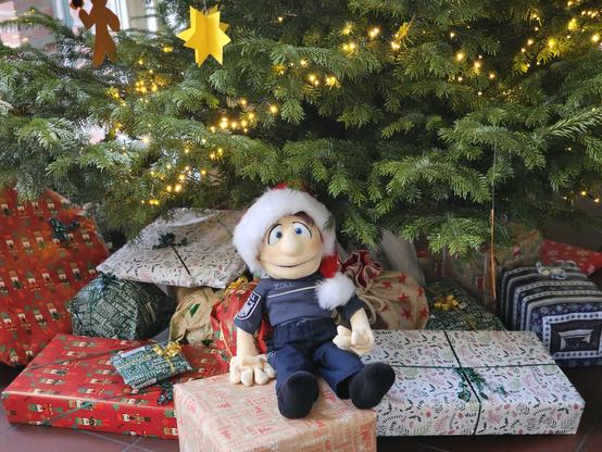 Eine Puppe mit Zoll-Dienstkleidung und Weihnachtsmütze sitzt auf Geschenken unter einem Weihnachtsbaum.